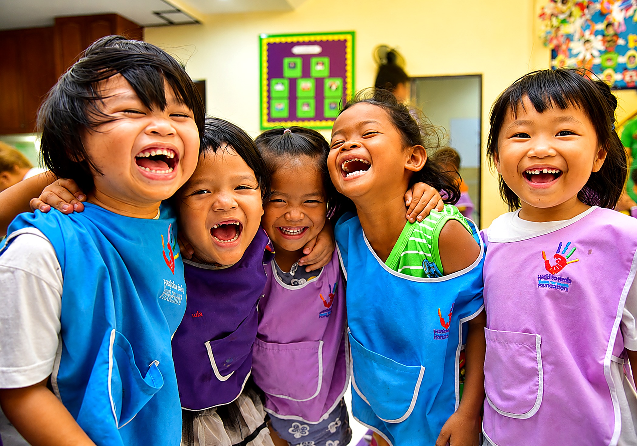 Five laughing children in Hand to Hand pre-school aprons, arms around each other
