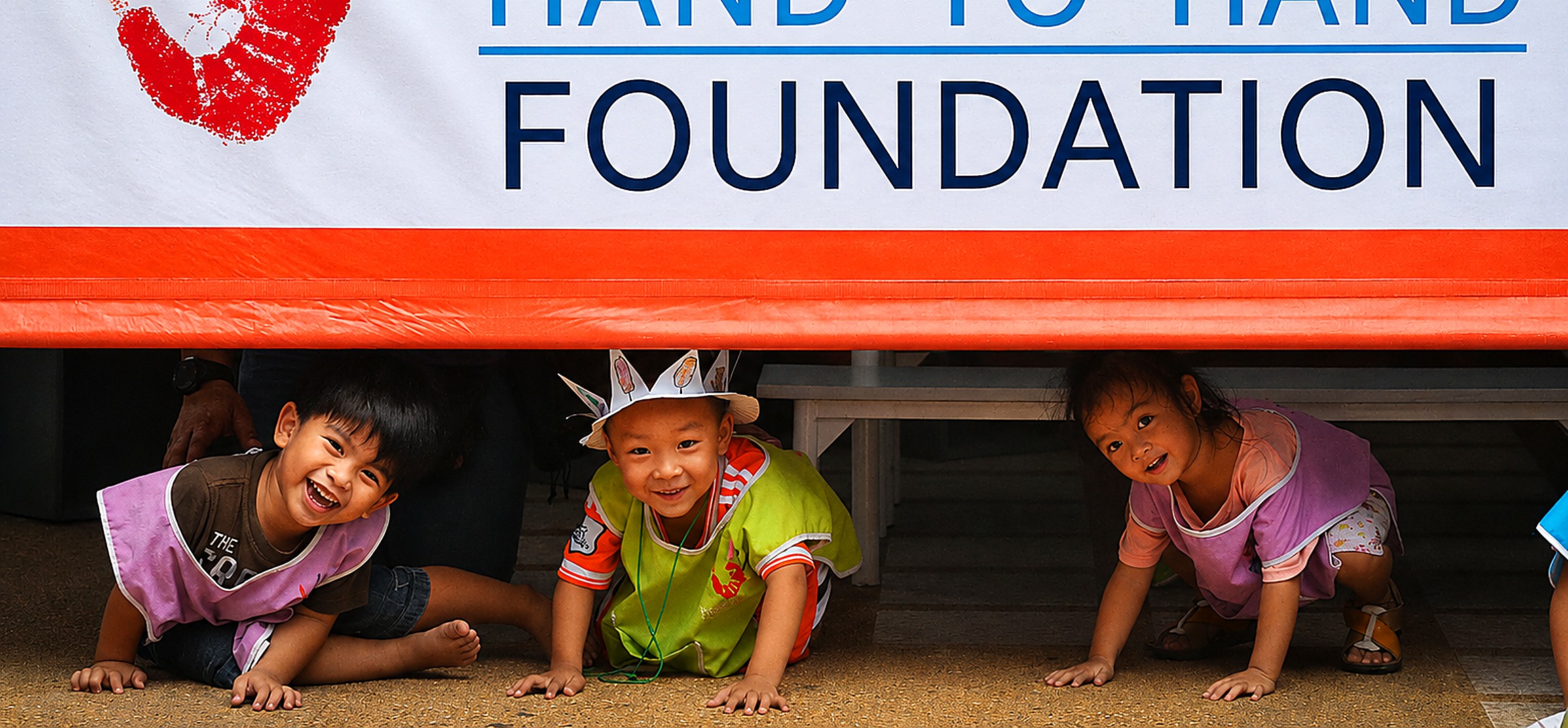 Three children laughing and playing beneath the Hand to Hand Foundation banner